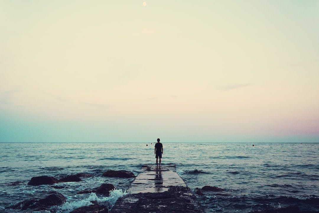 A man stands on a rocky path in the ocean facing the unknown.